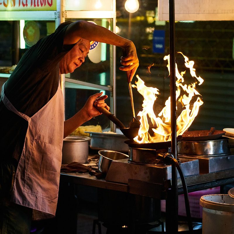 A street food cook tossing a wok over open flames at a night stall in Bangkok