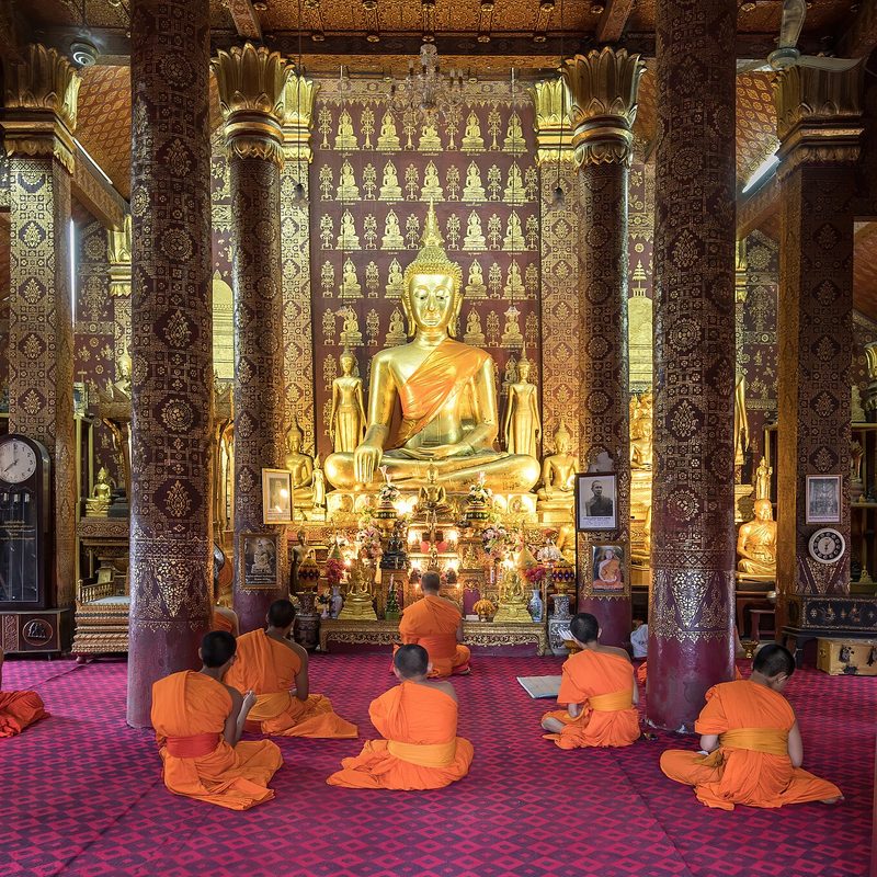 Monks in orange robes kneeling in prayer before a gilded Buddha inside a temple hall