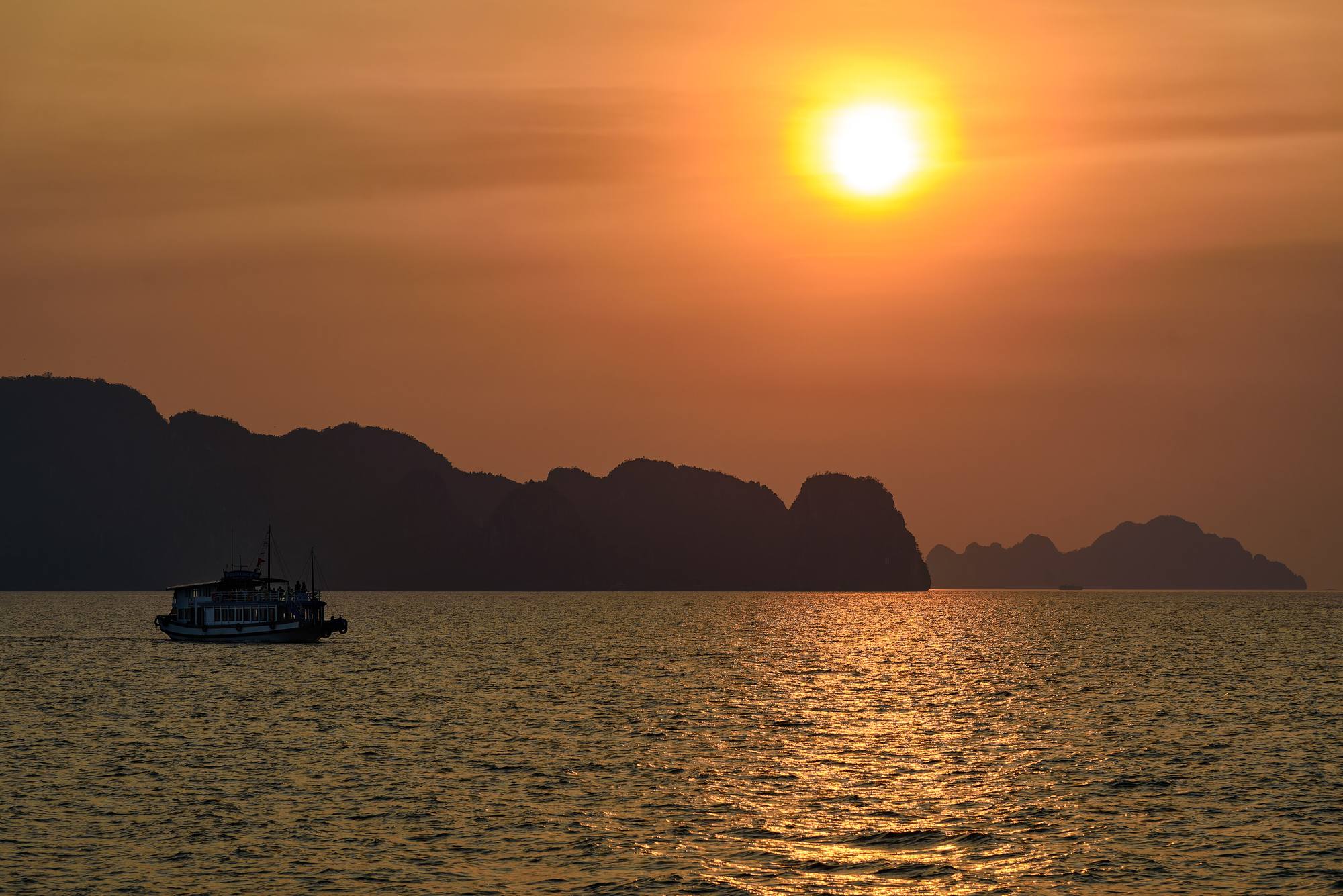 The sun setting behind the limestone islands of Ha Long Bay, a boat crossing the water below