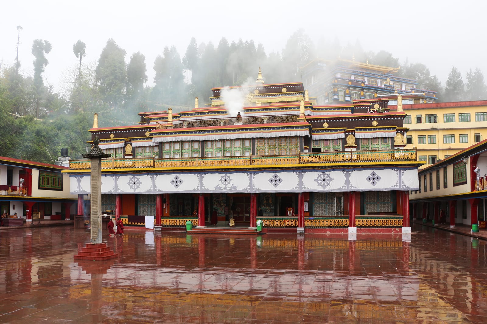 The painted facade of Rumtek Monastery reflected in its rain-wet courtyard, mist in the trees behind