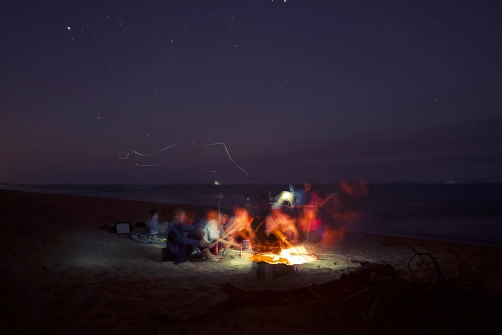People sitting around a bonfire on a dark beach under a sky full of stars