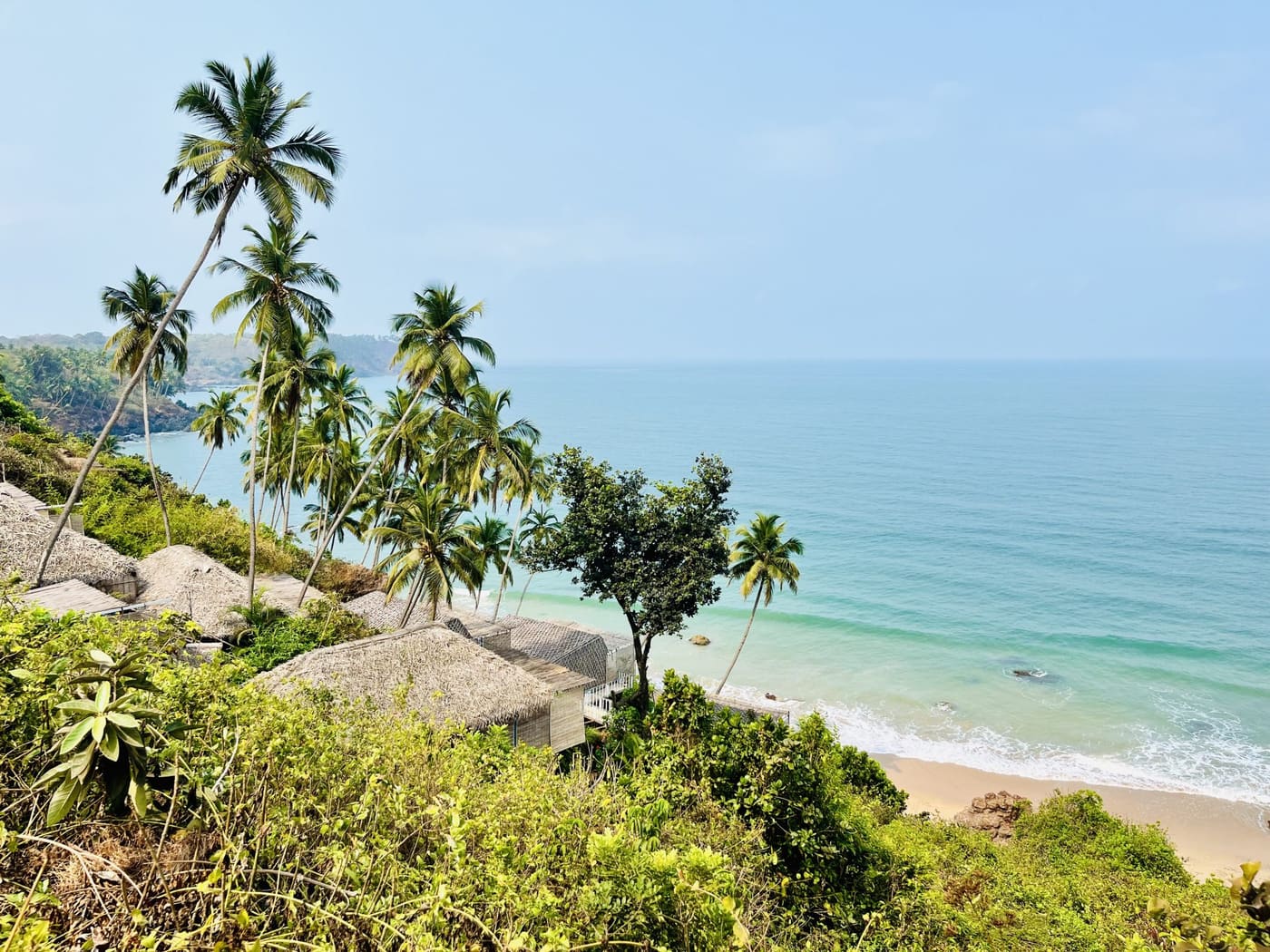 Coconut palms and thatched huts on a green headland above turquoise water in South Goa